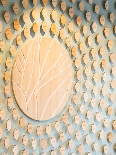 Foto der "Wall of Change": Wandinstallation mit vielen kleinen Holzblättern mit Nachhaltigkeitsmaßnahmen hängen an einer Wand, in der Mitte ein rundes Holzsymbol