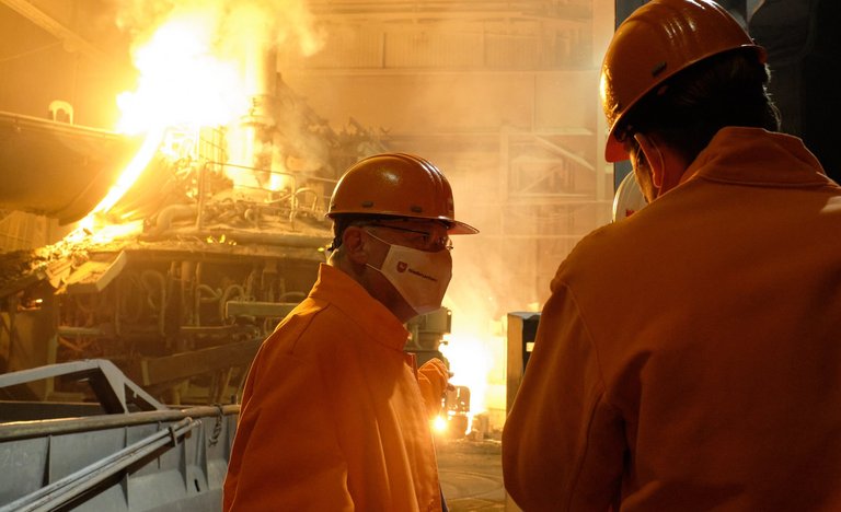 Mann mit Helm und Mund-Nasen-Maske im Gespräch mit anderen Mitarbeitenden. Im Hintergrund große Maschinen und ein Kessel mit gleißend hellem Stahl