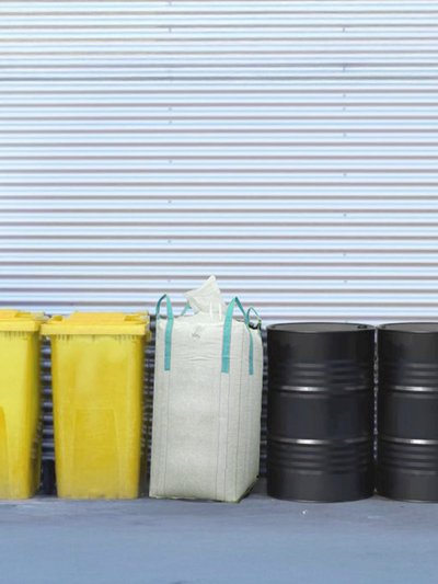Foto von mehreren gelben Tonnen, ein großer Sammelsack und schwarze Industriefässer zur Sammlung von Kunststoffrezyklaten stehen nebeneinander