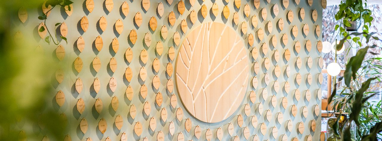 Foto der "Wall of Change": Wandinstallation mit vielen kleinen Holzblättern mit Nachhaltigkeitsmaßnahmen hängen an einer Wand, in der Mitte ein rundes Holzsymbol