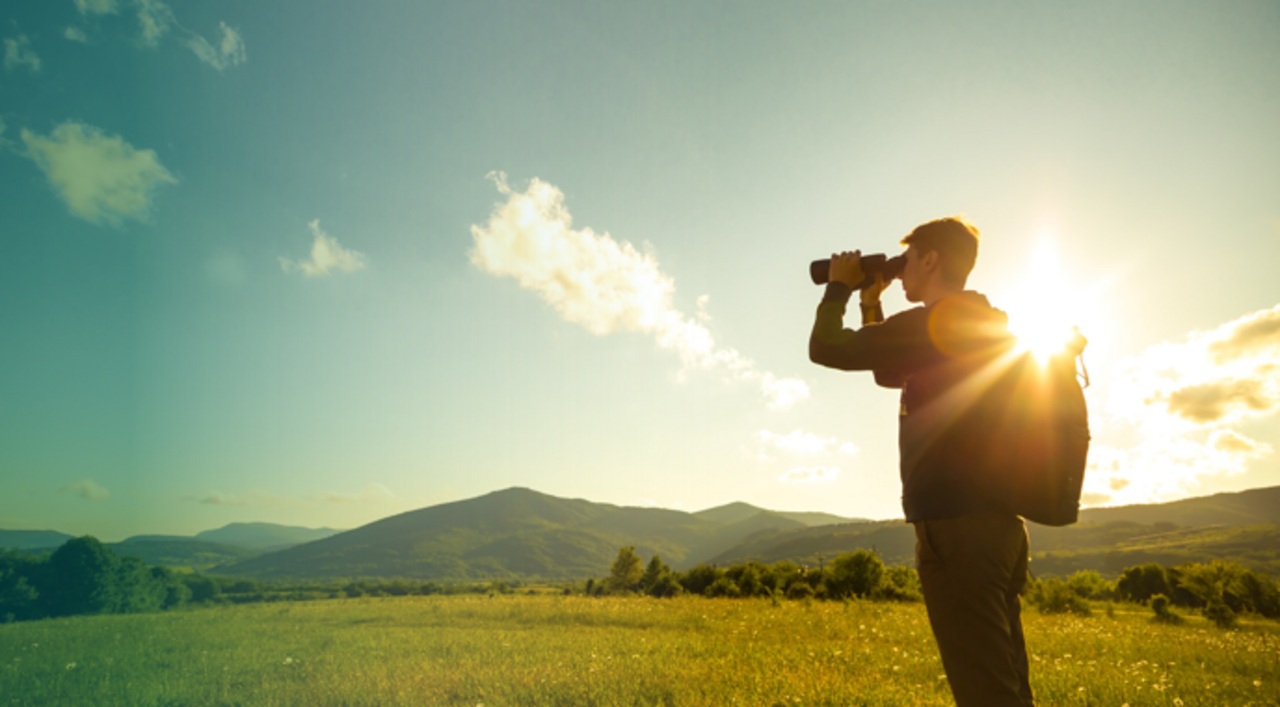 Bild von einem Mann, der im Sonnenschein auf einer Wiese steht und durch ein Fernglas sieht