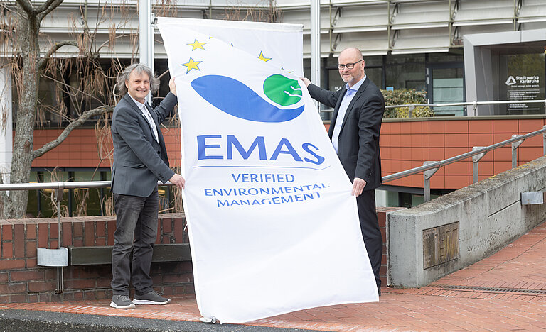 Zwei Männer stehen mit einer großen Flagge am Fahnenmast. Die Flagge ist weiß und trägt das EMAS-Logo.