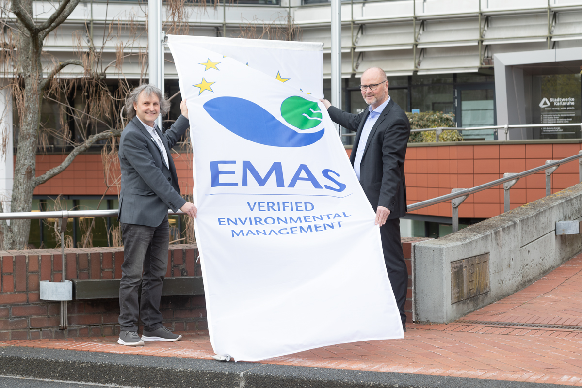 Zwei Männer stehen mit einer großen Flagge am Fahnenmast. Die Flagge ist weiß und trägt das EMAS-Logo.